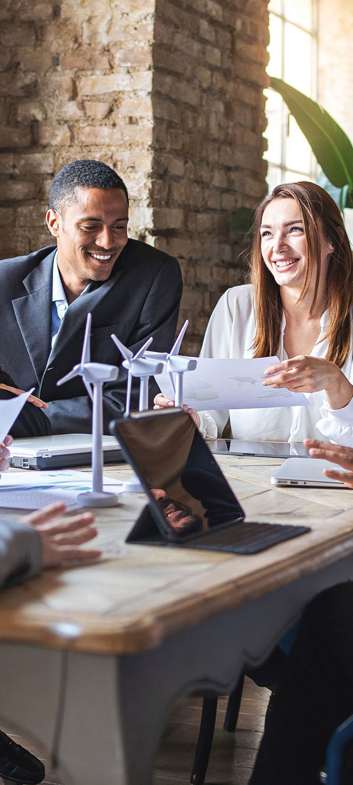 Professional people sit around a table having a meeting, there are papers and laptops on the table, one lady is is holding a piece of paper smiling and animatedly discussing something, a man to her left looks over at the paper smiling