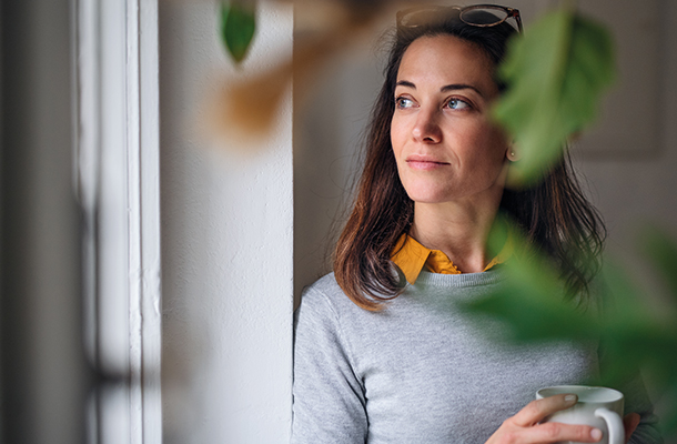 Lady in grey jumper holding a coffee cup looking wistfully out of a window with plants in the foreground