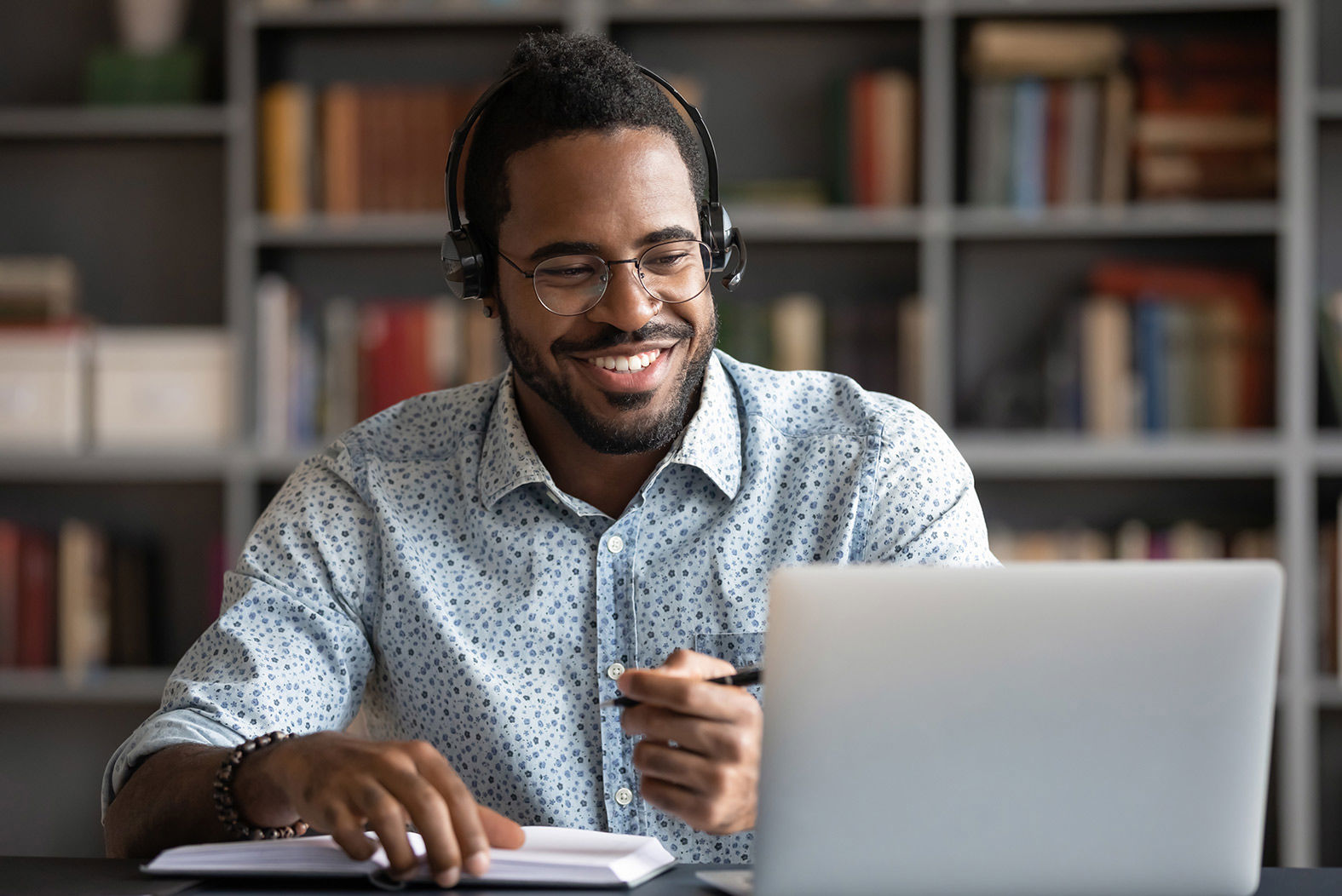 Smiling African man wearing headphones looking at laptop screen and writing in a pad of paper. He is wearing a blue dotted shirt. Blurred bookshelves are behind him