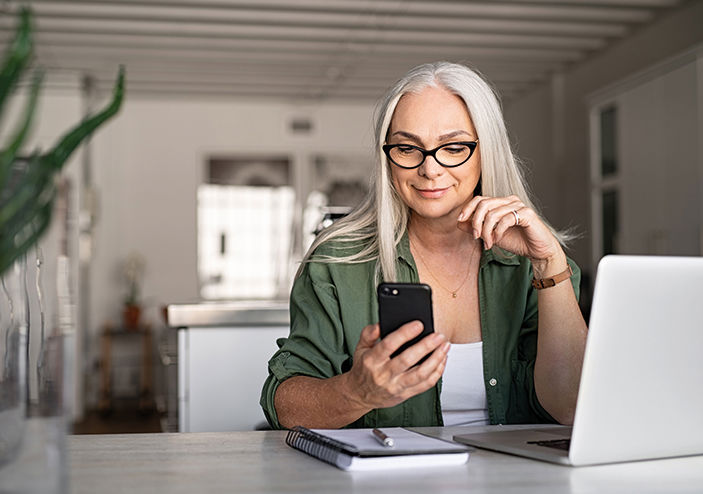 A lady with flowing white hair, sits at a desk smiling and looking at a phone in her hand. She has an open laptop next to her. She is wearing glasses and a green shirt. 