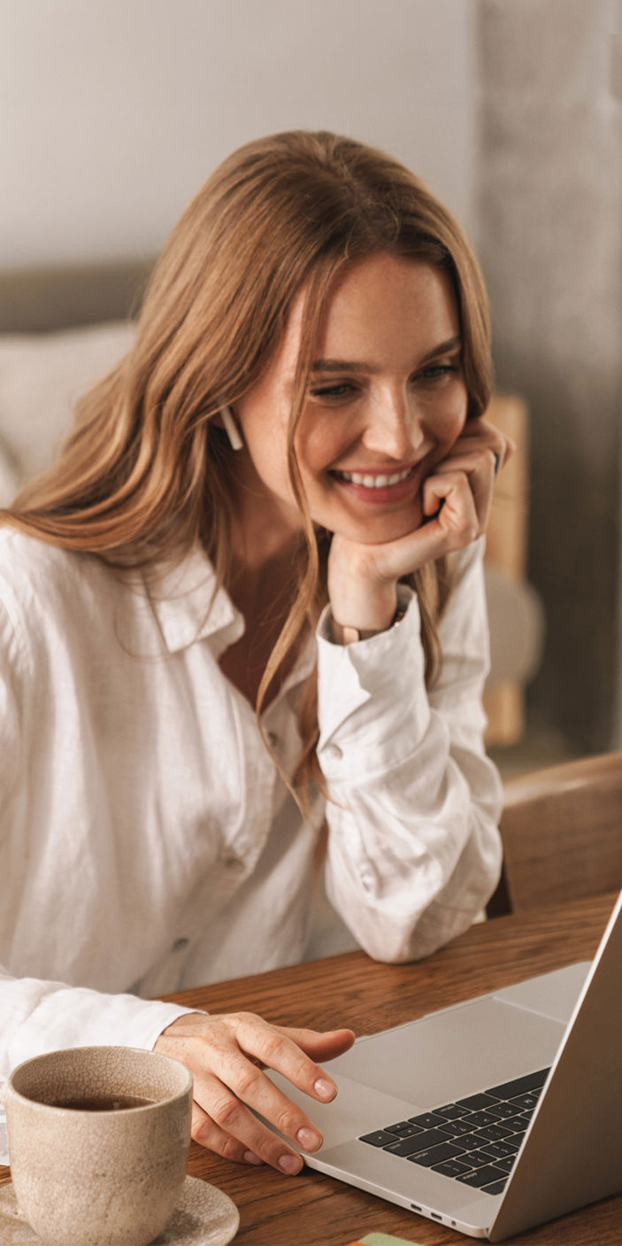 A lady in a white shirt smiles at the laptop on the table she is sitting at with a coffee cup to her side