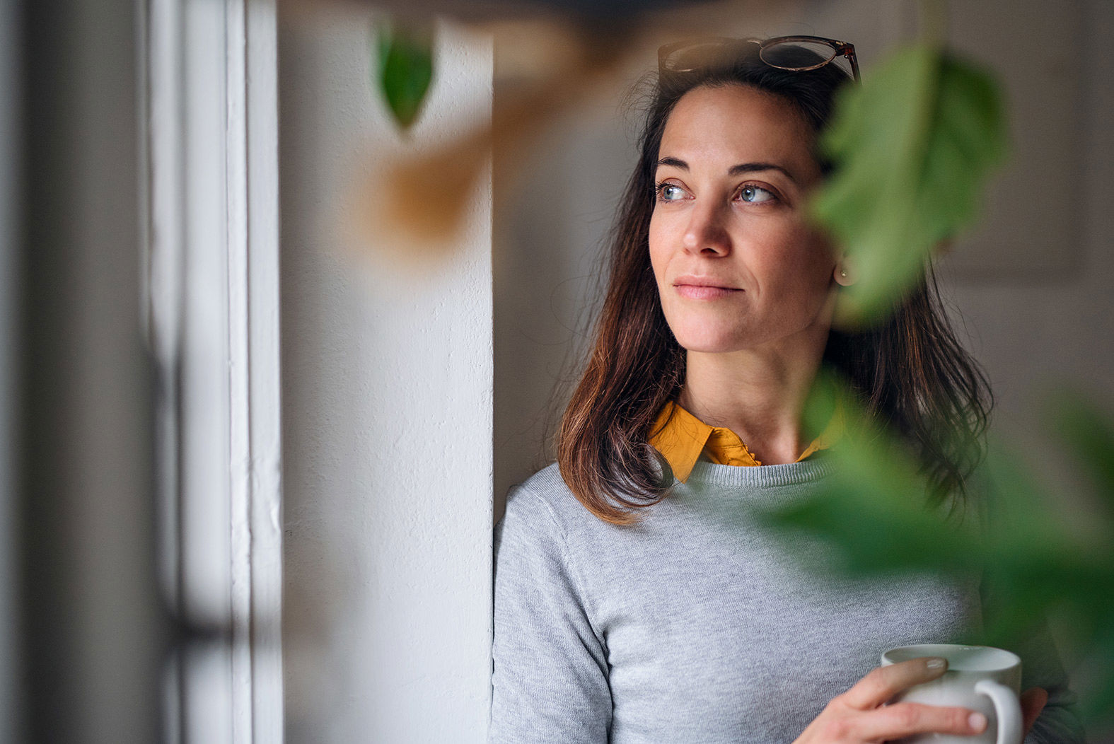 Lady in grey jumper holding a coffee cup looking wistfully out of a window with plants in the foreground