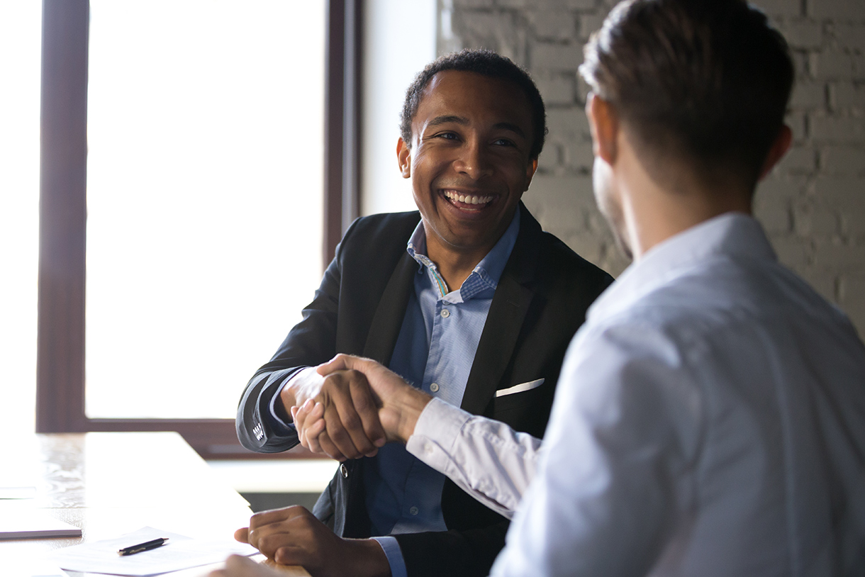 Two men sit at a desk, one facing the camera smiling, wearing a suit jacket and blue shirt, he is shaking the hand of another business professional slightly off camera also wearing a blue shirt