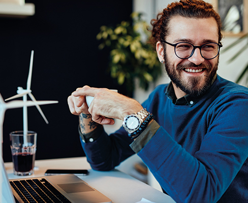 Man with beard and glasses looking to his right laughing at something off camera, he is holding a coffee cup and sitting at an open laptop